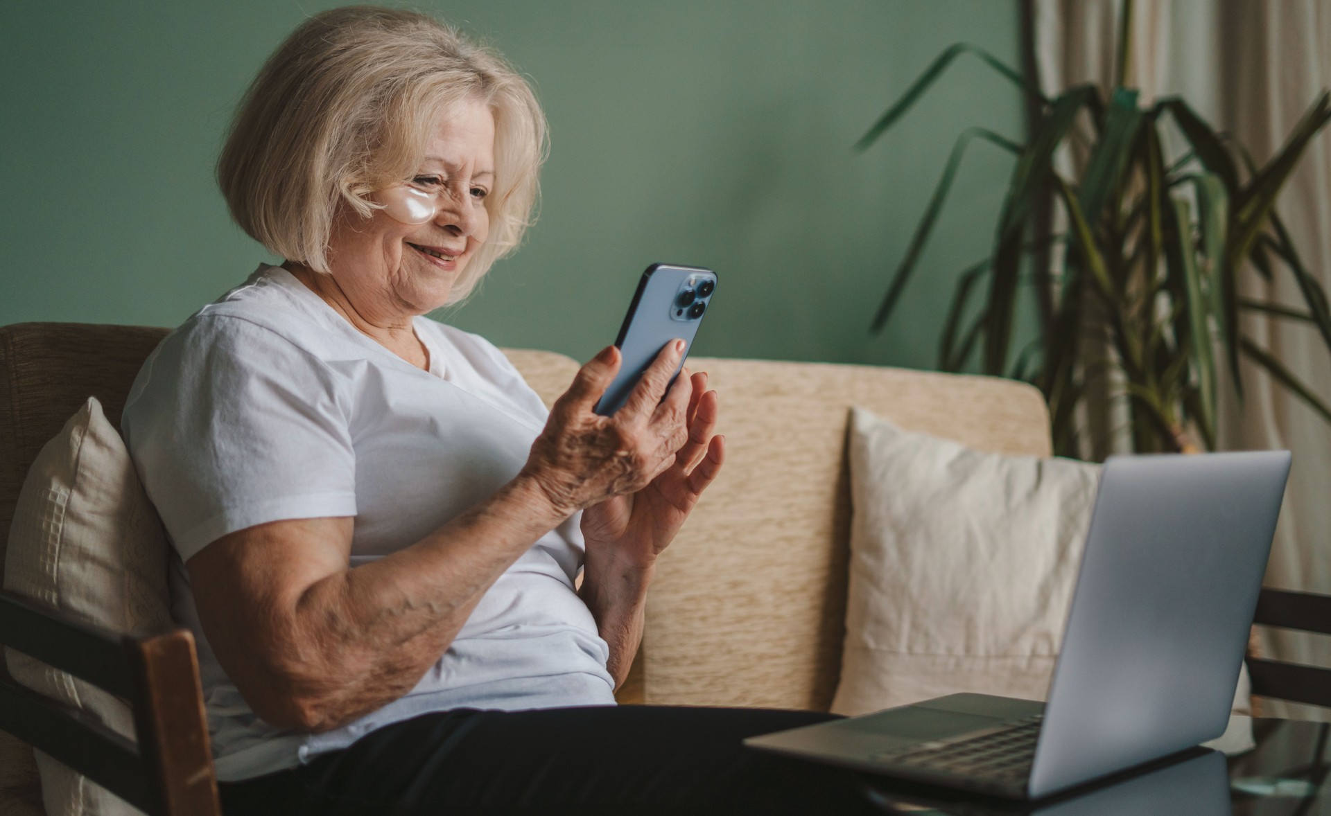 Femme mature avec des cache-yeux sur le visage assise sur le canapé devant un ordinateur portable et utilisant un téléphone portable. Communication par téléphone mobile.