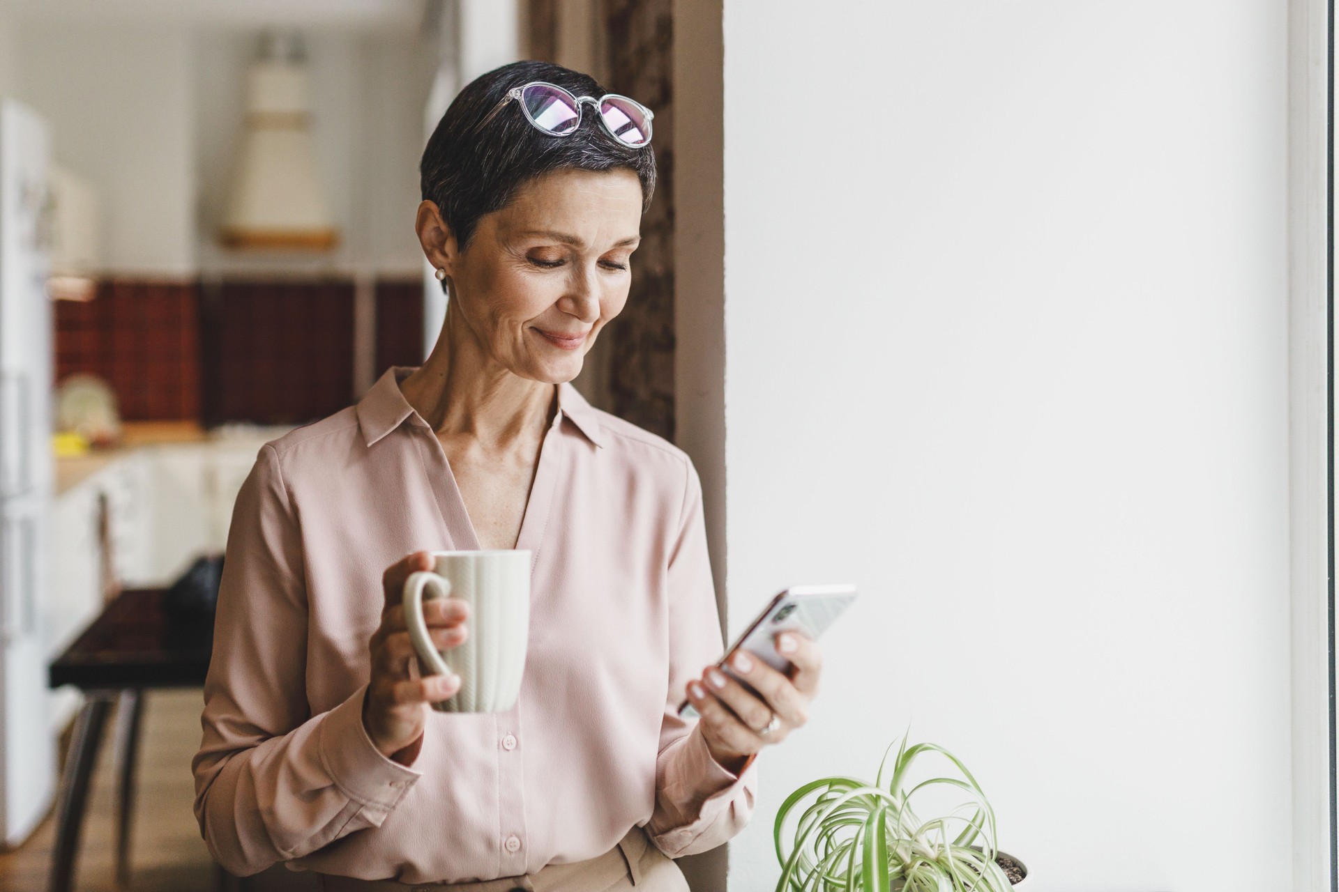 élégante femme mûre en chemise de soie élégante avec des lunettes sur la tête buvant du café et utilisant un smartphone, passant la matinée à la maison avant d’aller travailler au bureau, naviguant les mises à jour du fil d’actualités des m�